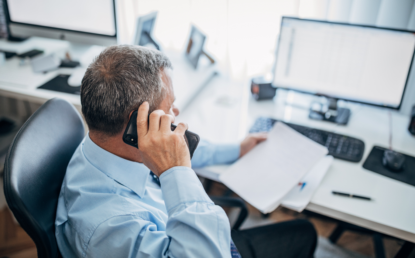 Værkfører sidder ved skrivebord på kontor og taler i telefon, mens han gennemgår dokumenter foran computer.